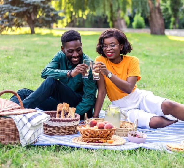 Photo of a young Black couple having a picnic together and smiling and clinking glasses together