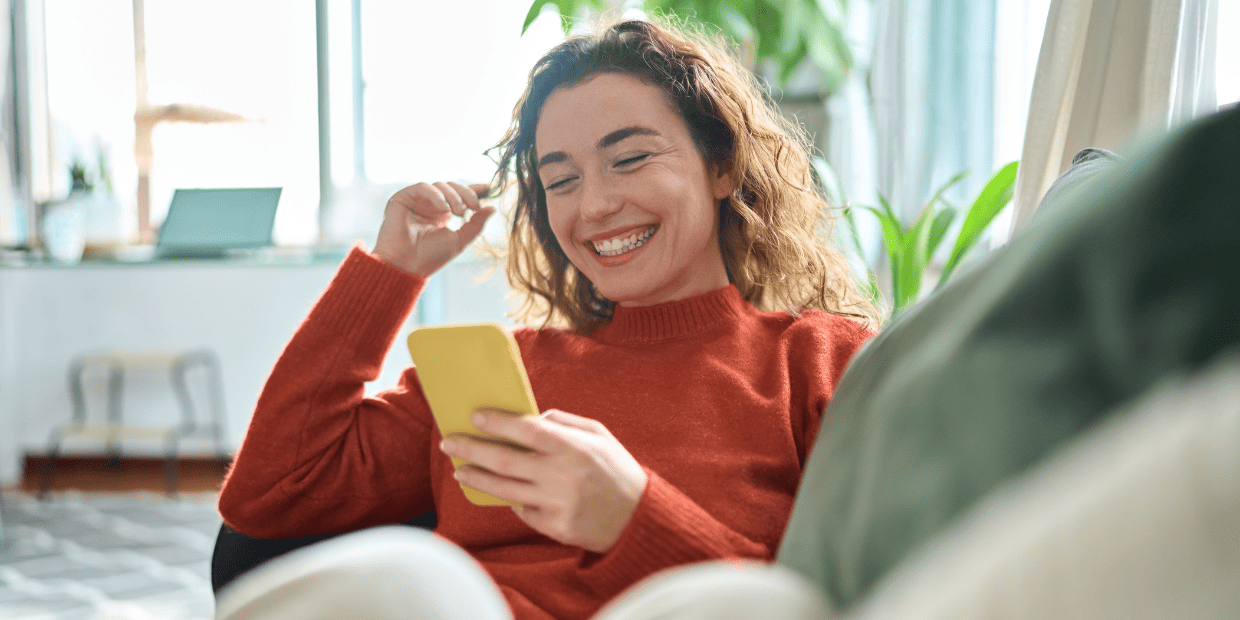 Young woman with curly hair lounging on couch and laughing at cellphone