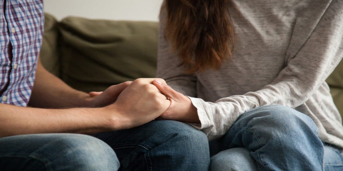 Photo of couple holding hands while sitting on a coach and having a serious conversation