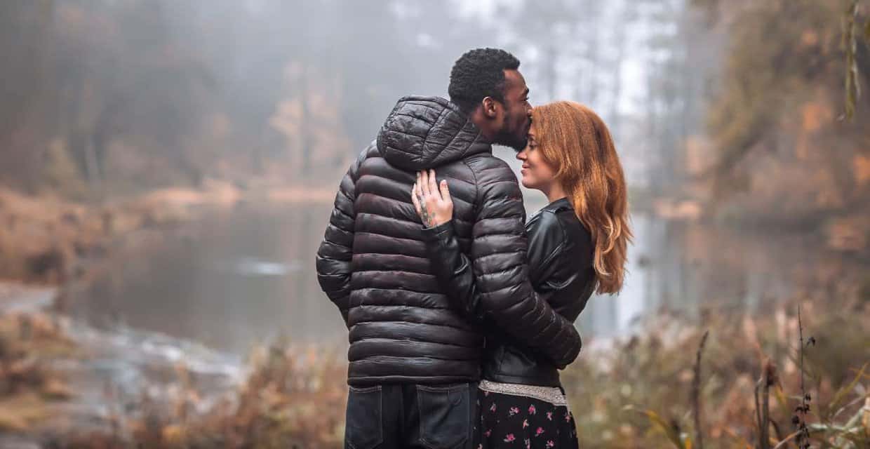 Photo of a Black man kissing a red haired woman on the head as they cuddle in a forest